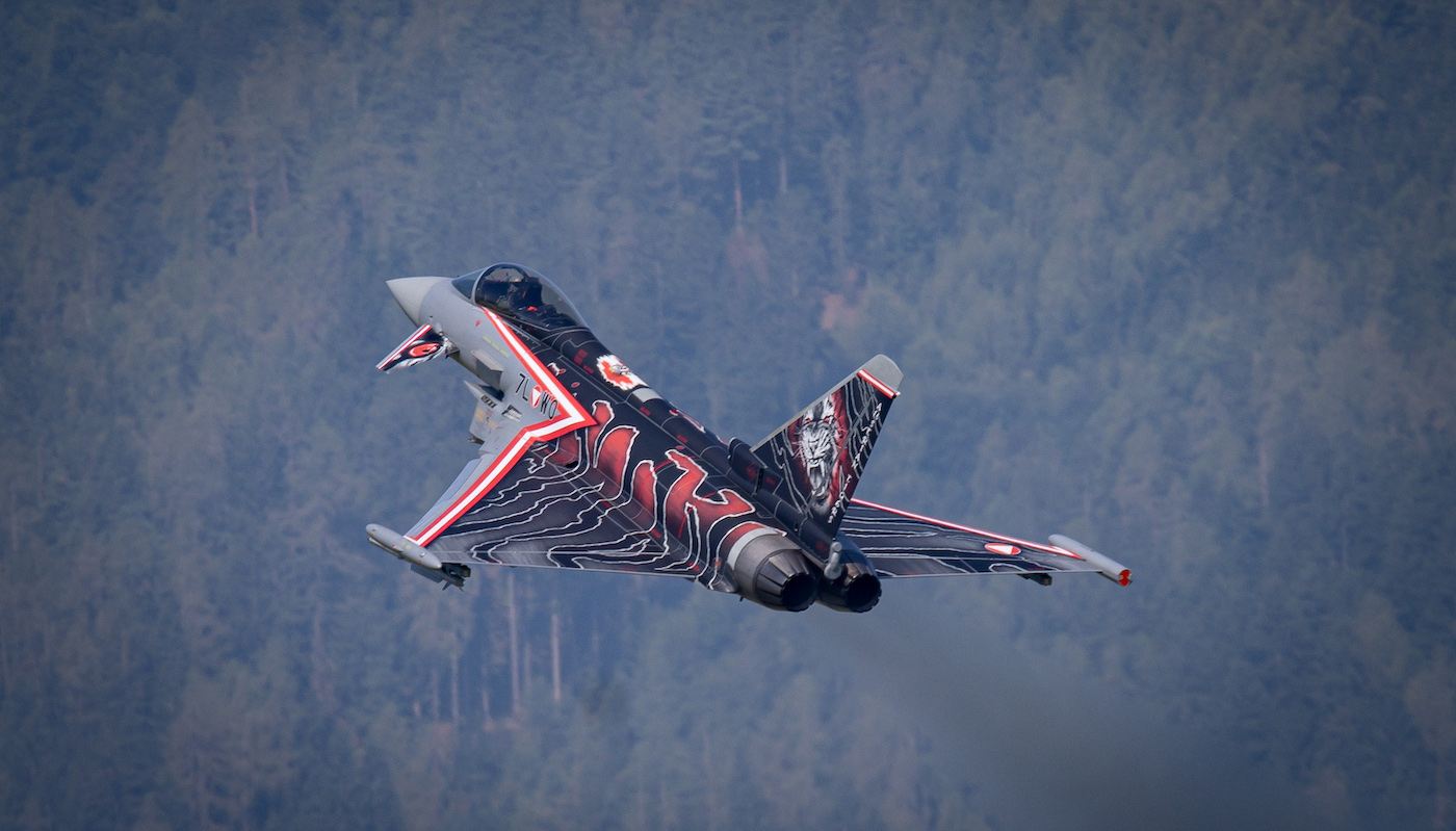 Ein dekorierter Kampfjet steigt mitten im Flug vor einem Hintergrund aus dichtem, nebligem Wald auf. Der Jet ist auf seinen Flügeln und seinem Heck mit kräftigen schwarz-rot-weißen Motiven verziert. Aus dem Auspuff des Heckteils strömt eine schwache Rauchfahne. Die Szene fängt den dynamischen und kraftvollen Aufstieg des Flugzeugs ein.