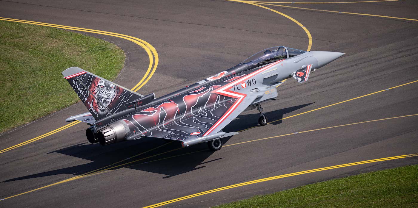 Ein Kampfjet mit einem dunklen, komplizierten Design in den Farben Rot und Schwarz und einem wilden Tigermotiv auf dem Heck rollt auf eine Startbahn. Das Flugzeug hat zwei Triebwerke und steht vor einem Hintergrund aus Gras und Asphalt mit markanten gelben Rollbahnmarkierungen.