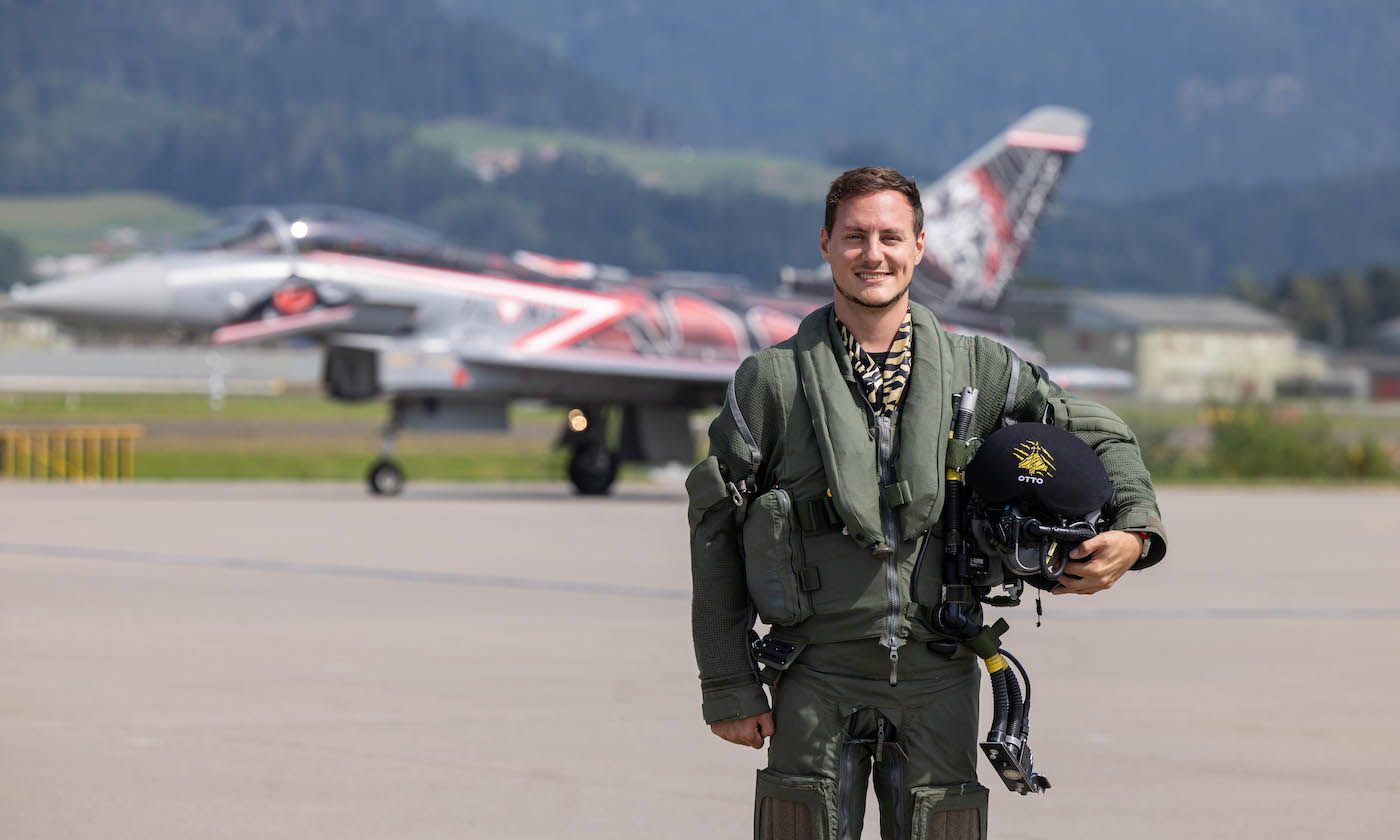 Ein Pilot steht auf einem Flugplatz vor einem Kampfjet mit deutlichen roten und schwarzen Markierungen. Der Pilot trägt einen grünen Fluganzug, hält einen Helm und lächelt in die Kamera. Im Hintergrund ist eine Berglandschaft mit grünen Feldern und Gebäuden in der Nähe des Flugplatzes zu sehen.