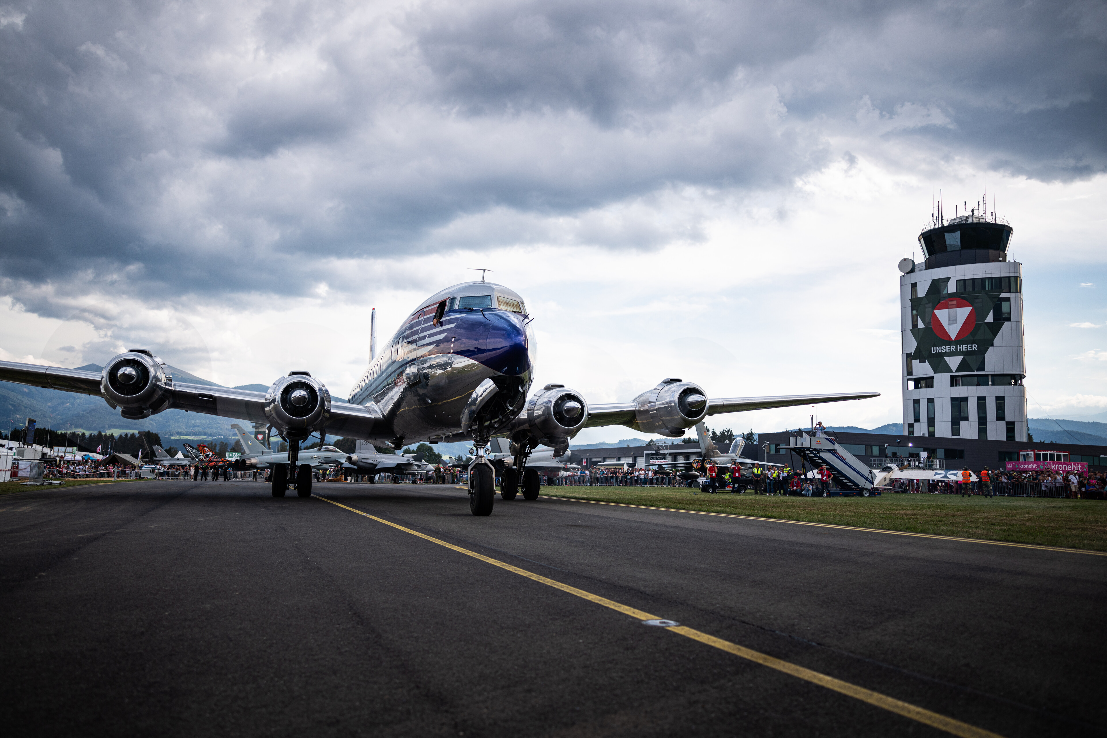 Ein altes Propellerflugzeug auf einer Landebahn in der Nähe eines Flugsicherungsturms mit einem bewölkten Himmel im Hintergrund.