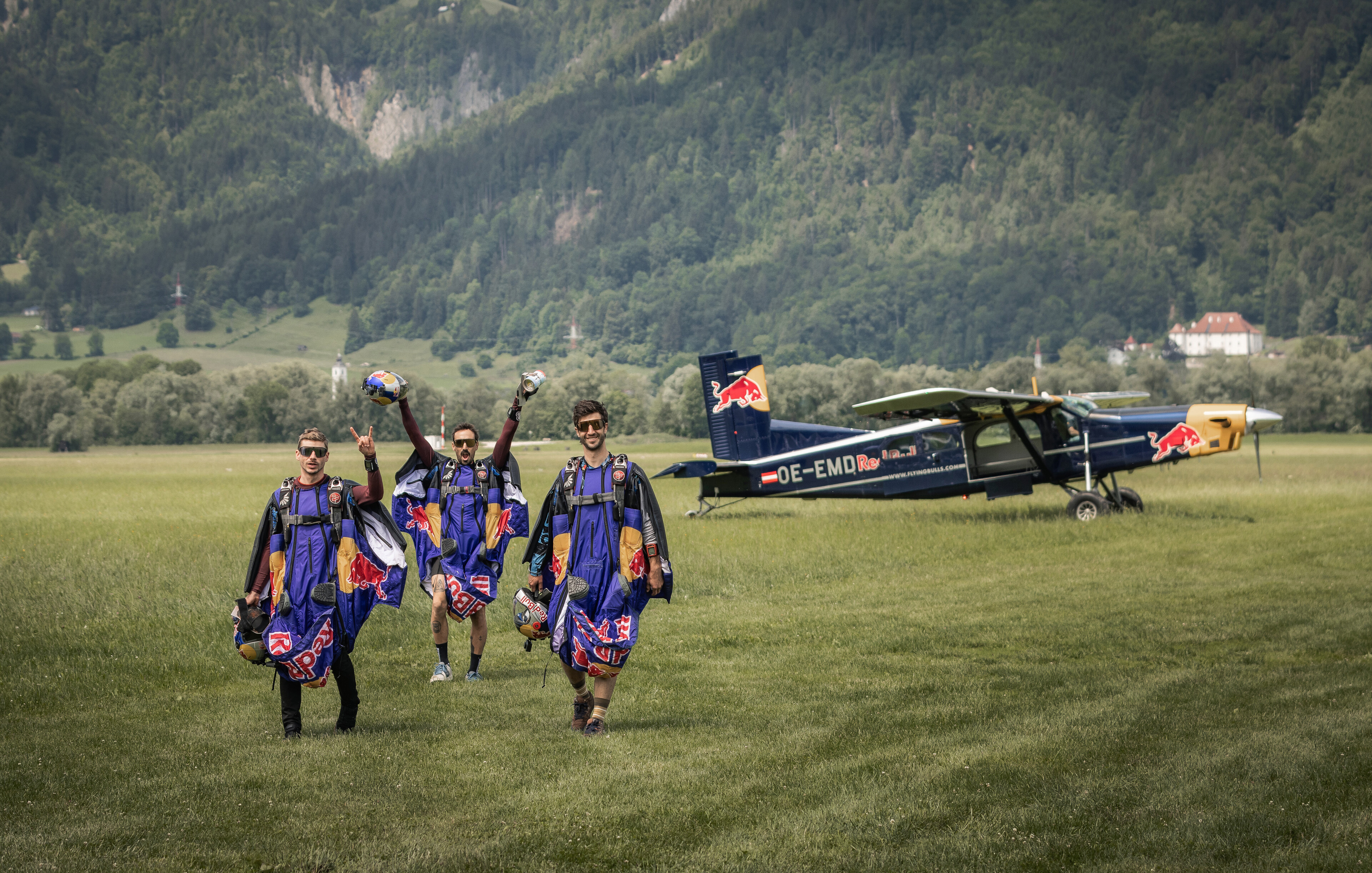 Drei Menschen in Wingsuits gehen von einem geparkten Red Bull-Flugzeug auf einer Wiese mit Bergen im Hintergrund weg.