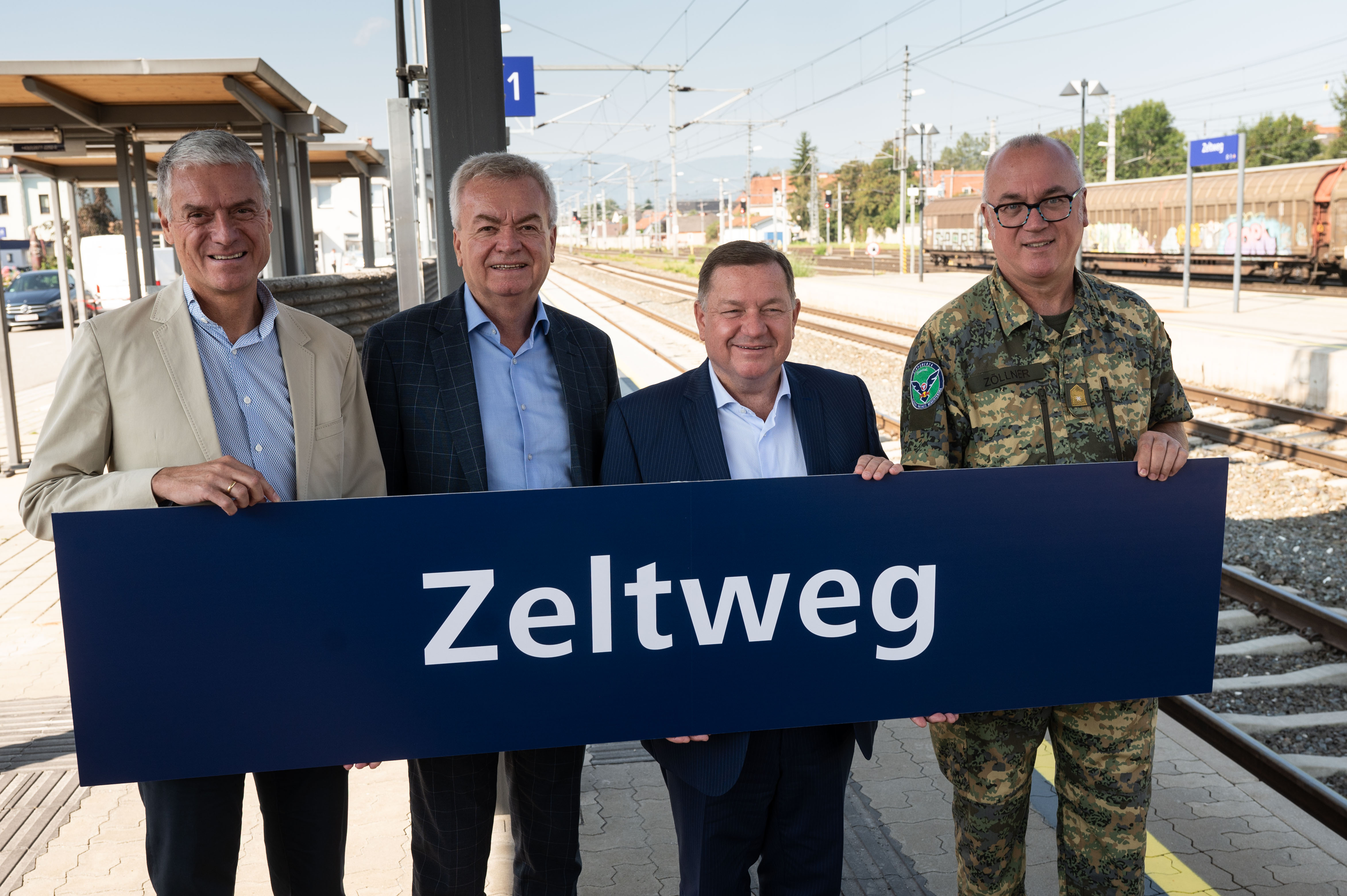 Vier Männer stehen auf einem Bahnsteig und halten ein Schild mit der Aufschrift „Zeltweg“, im Hintergrund sind Bahngleise und ein Gebäude zu sehen.