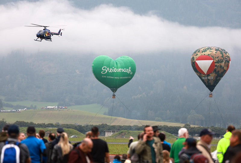 A helicopter and two hot air balloons, one green and heart-shaped, fly above a crowd in a hilly landscape.