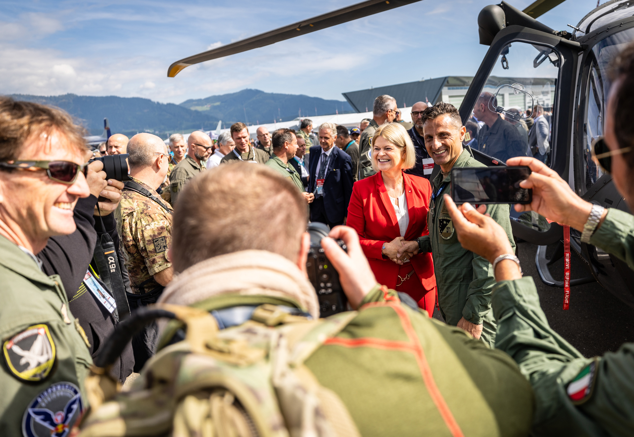 Eine Gruppe Militärangehöriger macht Fotos und trifft an einem sonnigen Tag in der Nähe eines Hubschraubers eine Frau im roten Mantel.