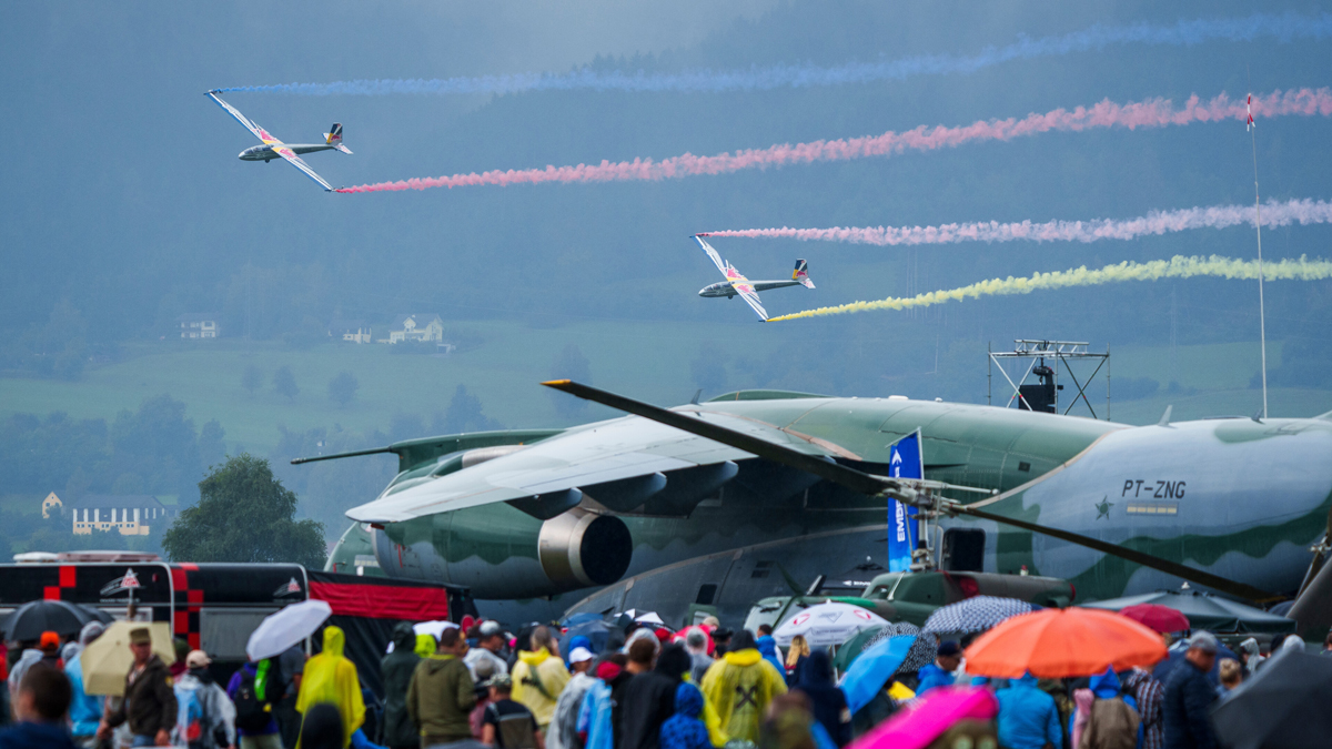 Flugschau mit zwei Flugzeugen, die farbige Rauchspuren über einer Menschenmenge hinterlassen, einem großen Militärflugzeug und Regenschirmen.