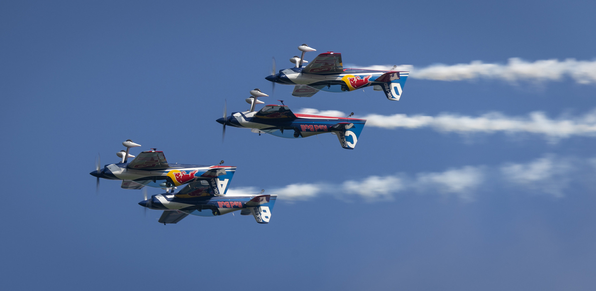 Vier Kunstflugzeuge mit Red Bull-Logos führen vor einem klaren blauen Himmel eine synchronisierte Flugroutine vor.