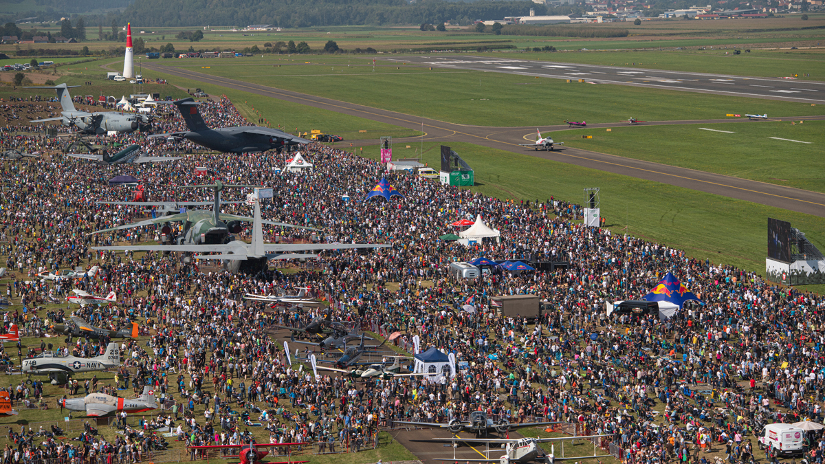 Große Menschenmenge besucht an einem klaren Tag eine Flugschau mit zahlreichen Militär- und Zivilflugzeugen, die in der Nähe einer Landebahn ausgestellt sind.