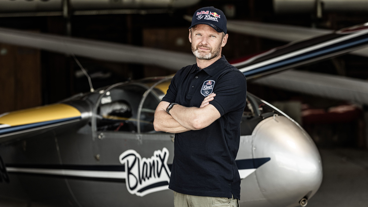 Mann in schwarzem Poloshirt und Mütze steht mit verschränkten Armen vor einem kleinen Flugzeug in einem Hangar.