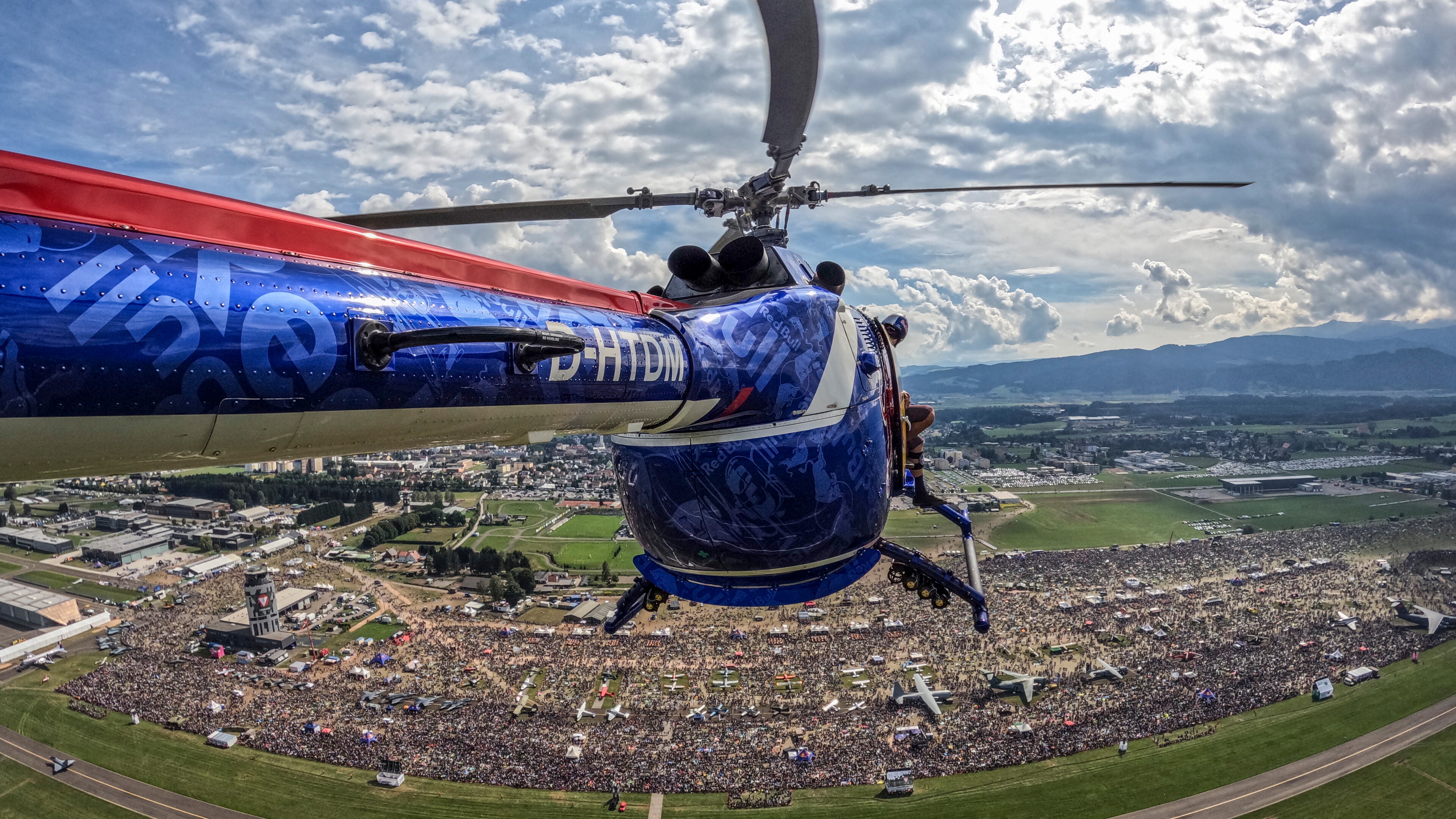 Ein Hubschrauber schwebt über einer riesigen Freiluftveranstaltung mit einer großen Menschenmenge, umgeben von einer Landschaft aus Feldern und Bergen in der Ferne.