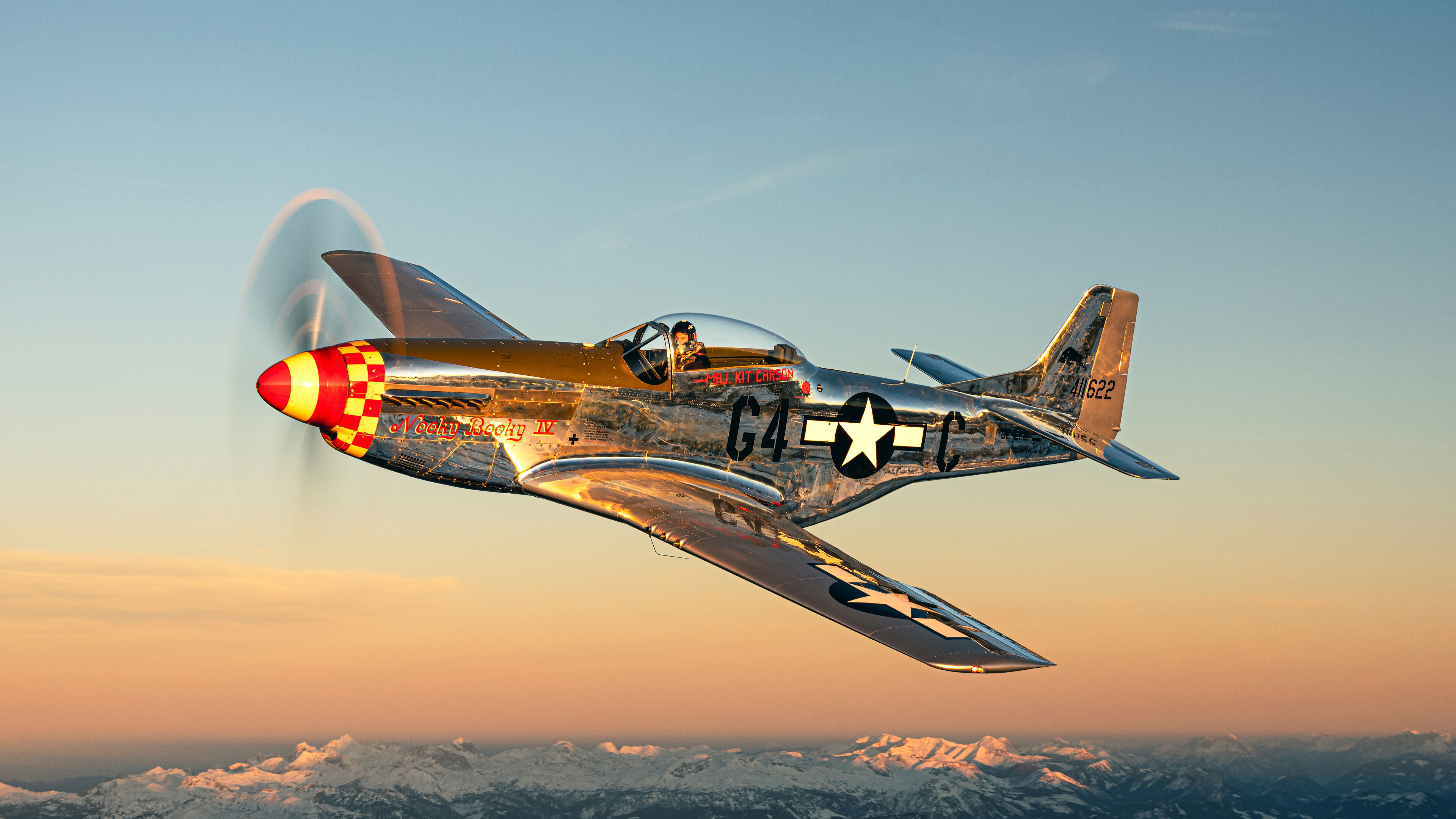 Ein silbernes P-51 Mustang-Kampfflugzeug aus dem Zweiten Weltkrieg im Flug, mit einem Sonnenuntergang im Hintergrund und schneebedeckten Bergen darunter.