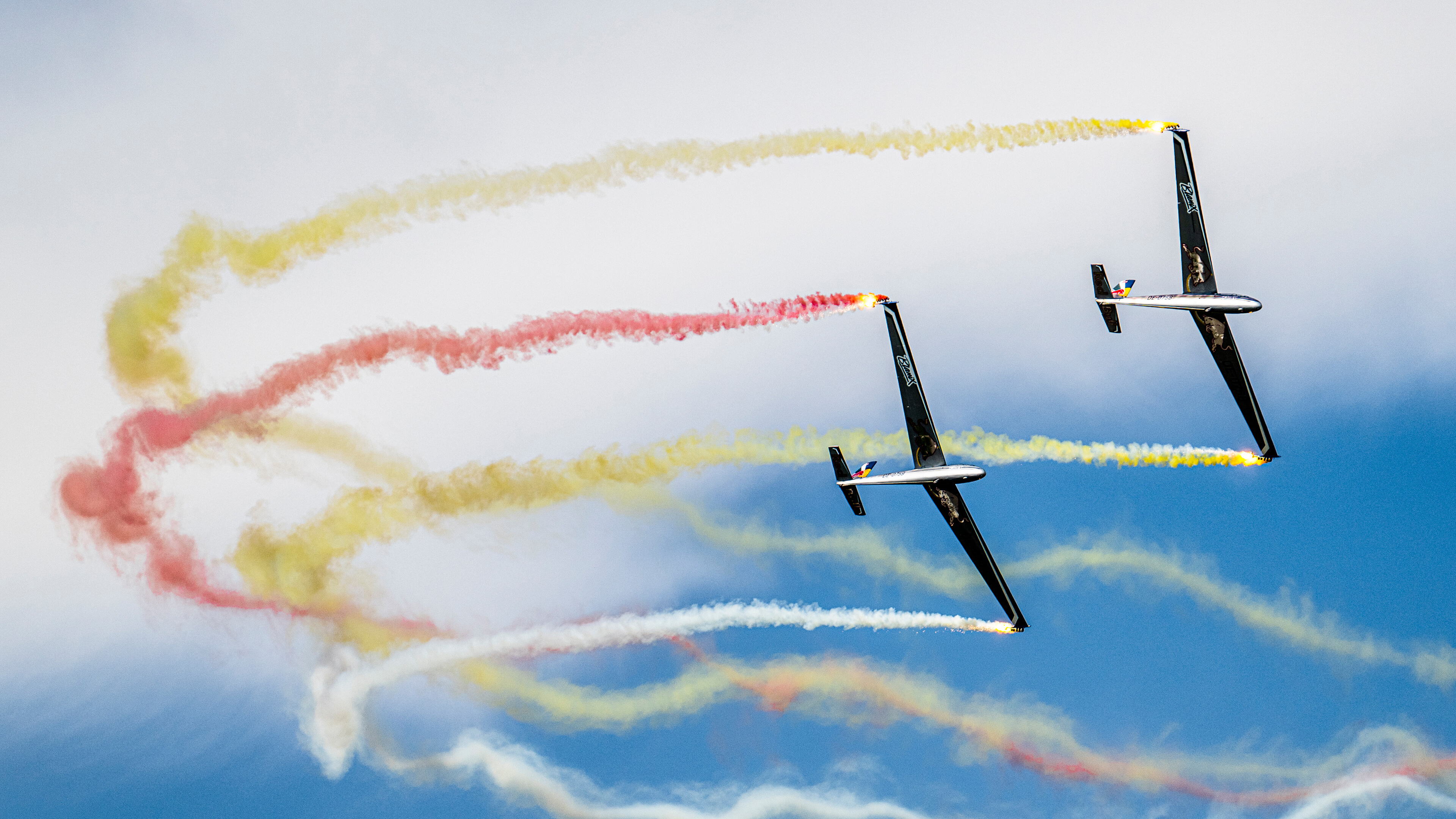 Zwei Flugzeuge führen Kunstflugmanöver aus und hinterlassen vor einem blauen Himmel eine bunte Rauchfahne in Rot, Gelb und Weiß.