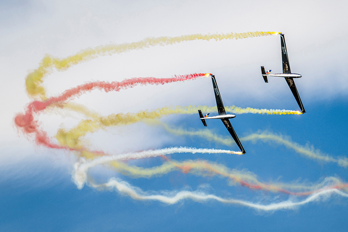 Two planes perform aerobatics with red, yellow, and white smoke trails against a blue sky.