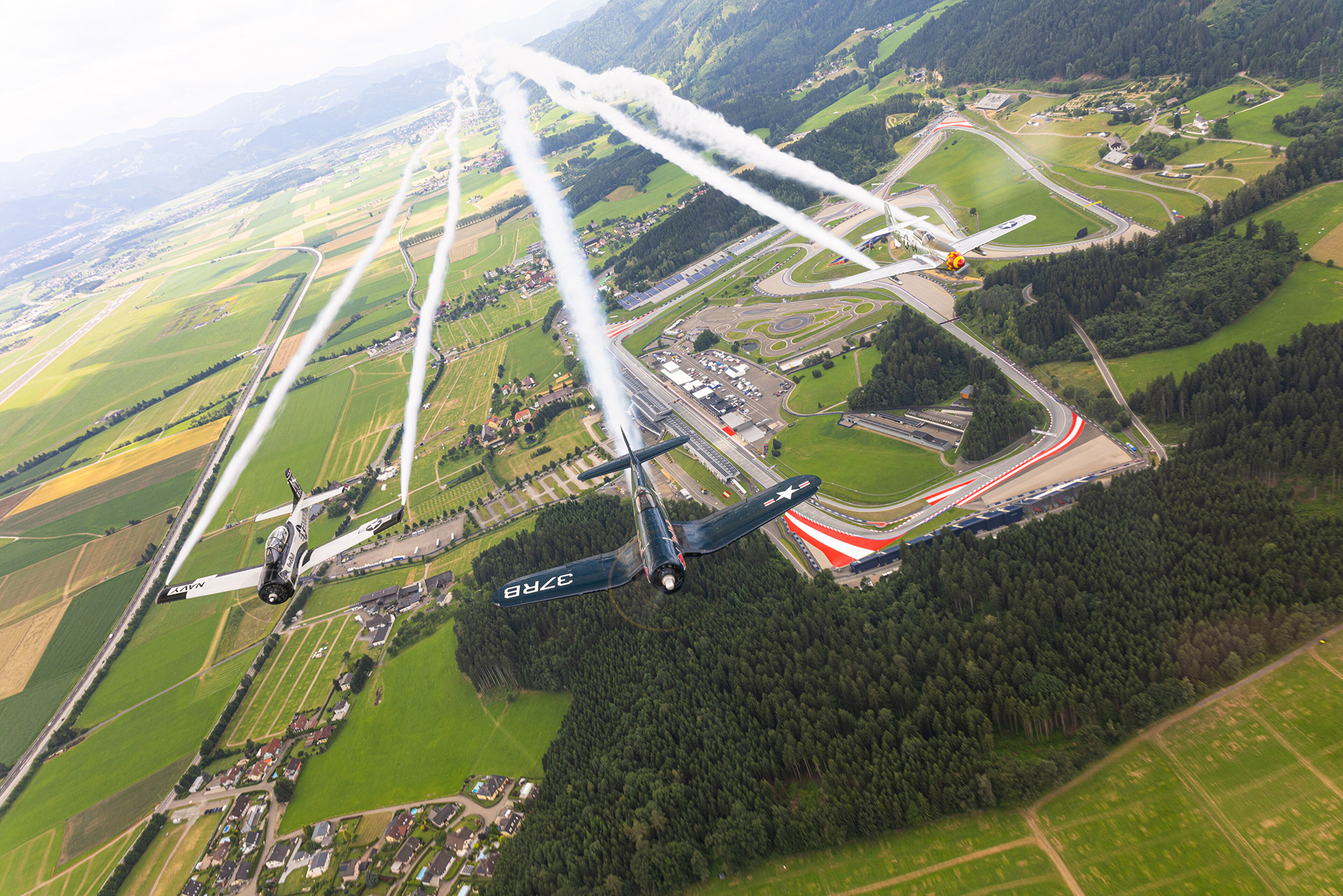 Three vintage fighter planes performing an aerial display over a scenic landscape with fields and a race track below.