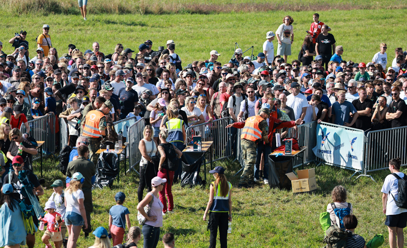 A crowd of people gathered outdoors, separated by metal barriers, with event staff in high-visibility vests.