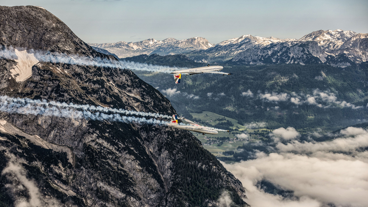 Zwei Flugzeuge führen ein akrobatisches Flugmanöver in der Nähe eines Berggipfels mit Rauchfahnen und einer malerischen Landschaft darunter durch.