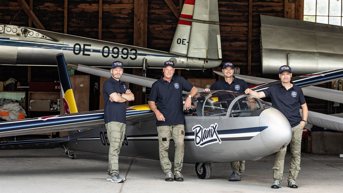 Vier Personen in passender Kleidung stehen neben einem Segelflugzeug in einem Hangar. Im Hintergrund sind weitere Flugzeuge zu sehen.